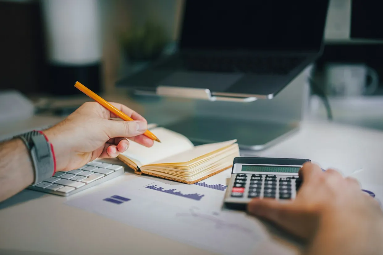 Working through admin figures with a notebook, calculator and printed charts beside a laptop