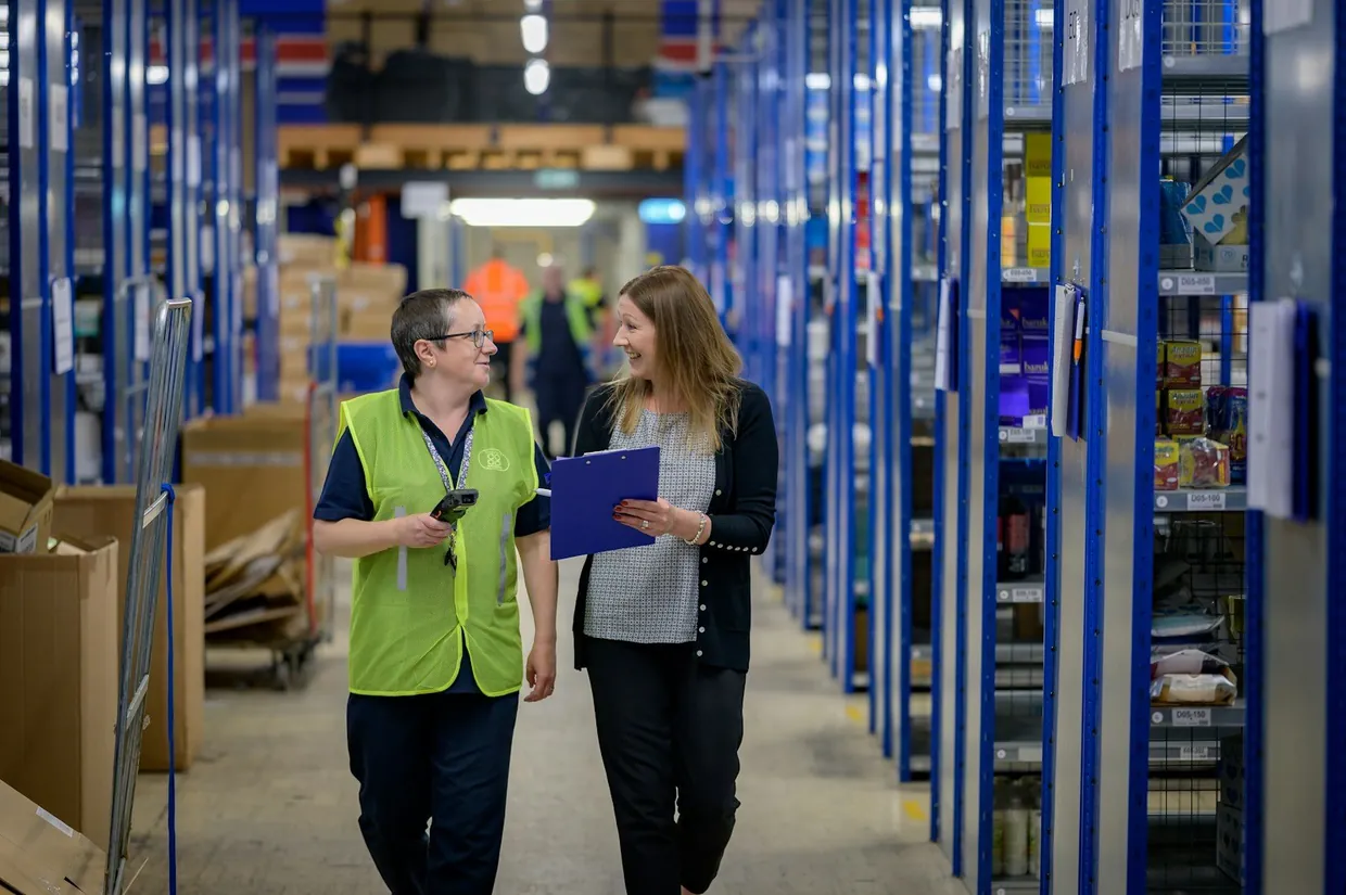 Warehouse staff with a handheld scanner and clipboard checking stock along shelving aisles