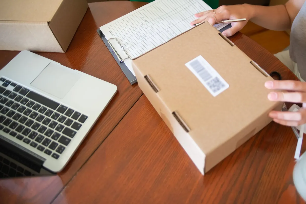 Hands checking a returned parcel and its barcode label against a clipboard record beside a laptop