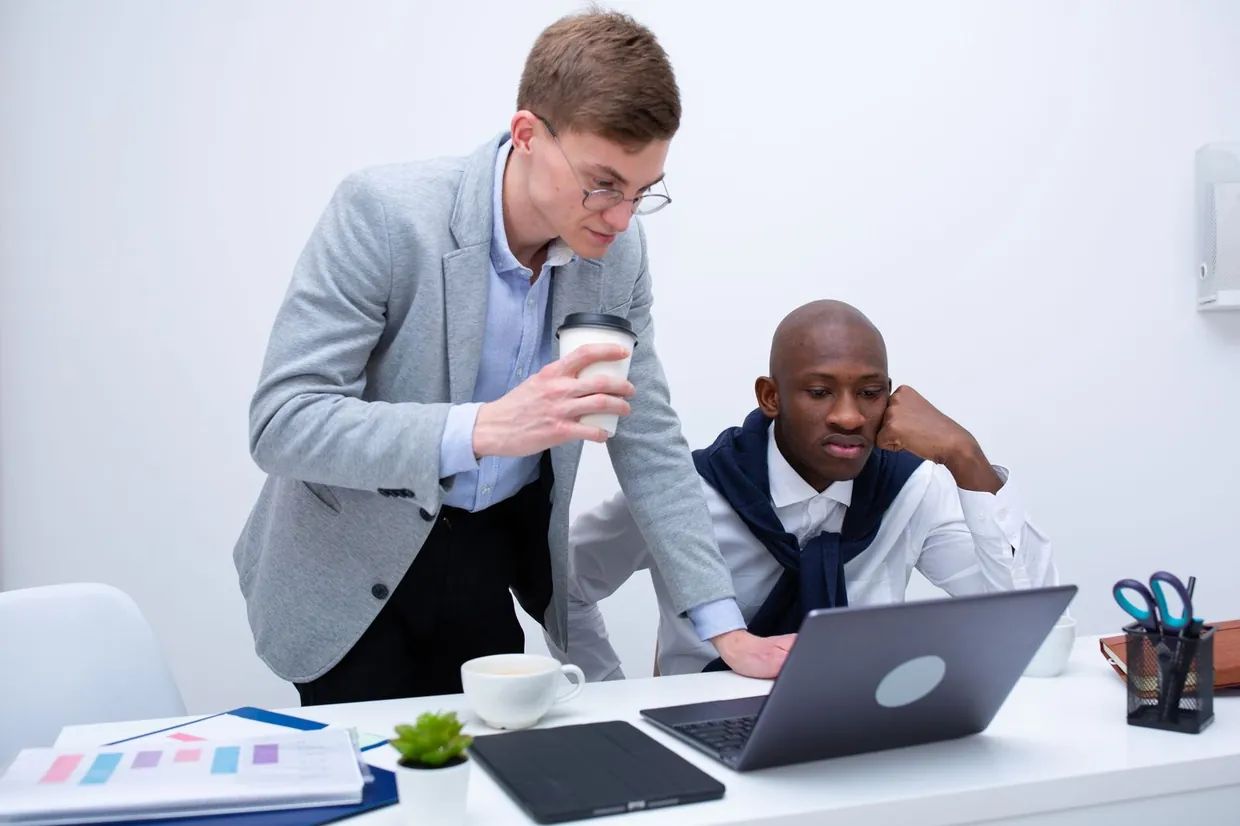 A colleague leans over a desk to walk a teammate through a task on a laptop