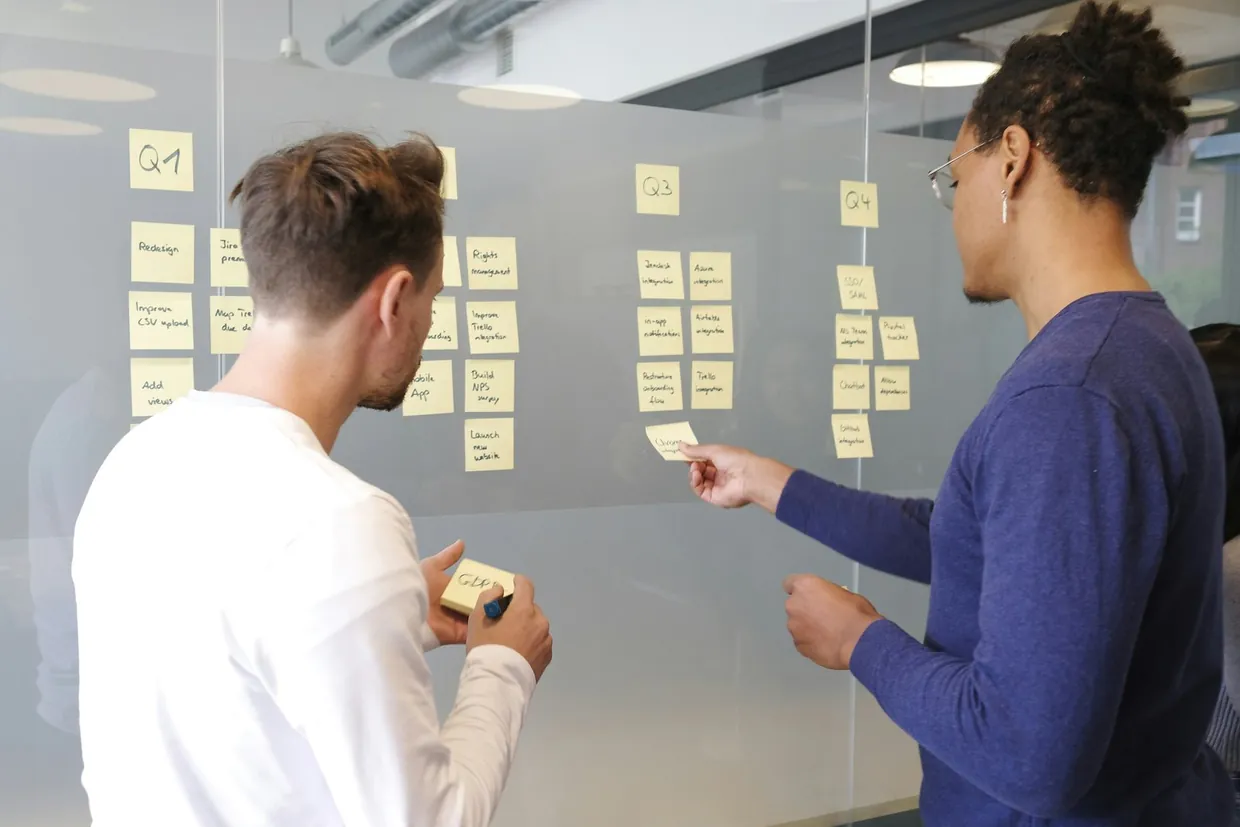 Two colleagues planning a help desk queue on a glass wall of sticky notes