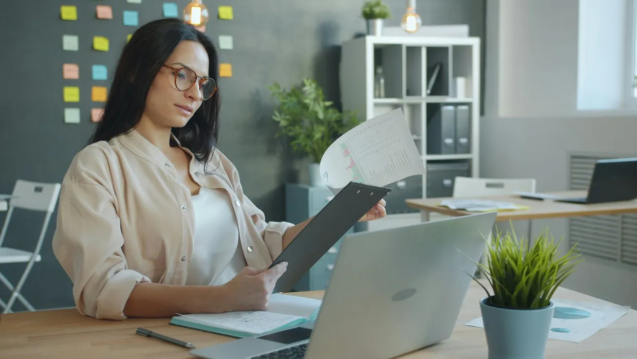 Assistant reviewing a printed plan on a clipboard against her laptop to weigh up admin hours