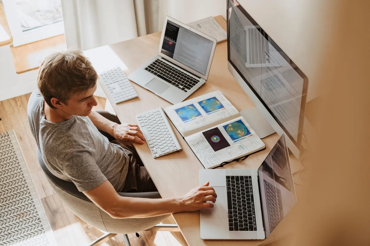 Man working across a laptop and second screen with a notebook beside him, keeping on top of email