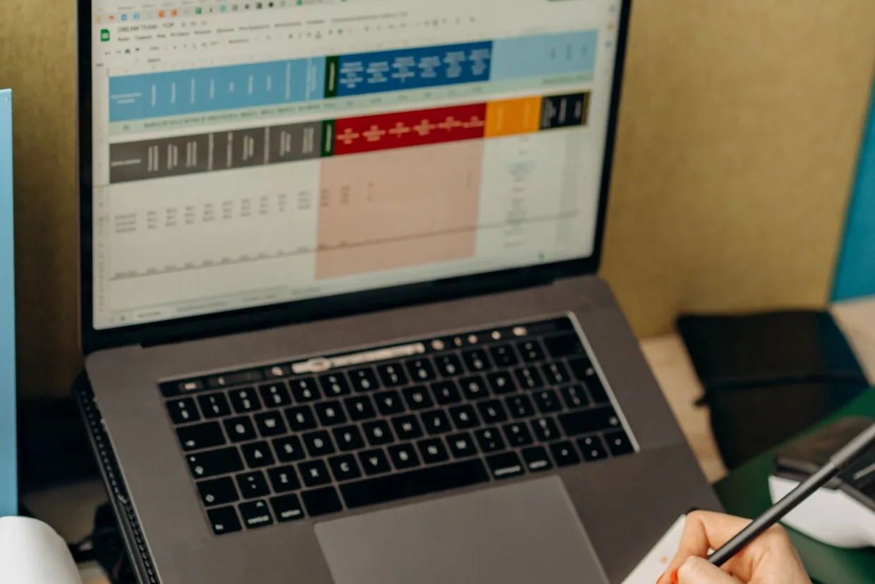 Hand holding a pen beside a laptop showing a colour-coded sales spreadsheet while working through a stock forecast