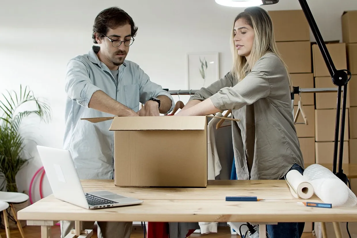 Two people packing an order into a cardboard box at a dispatch table, with tape and a laptop nearby