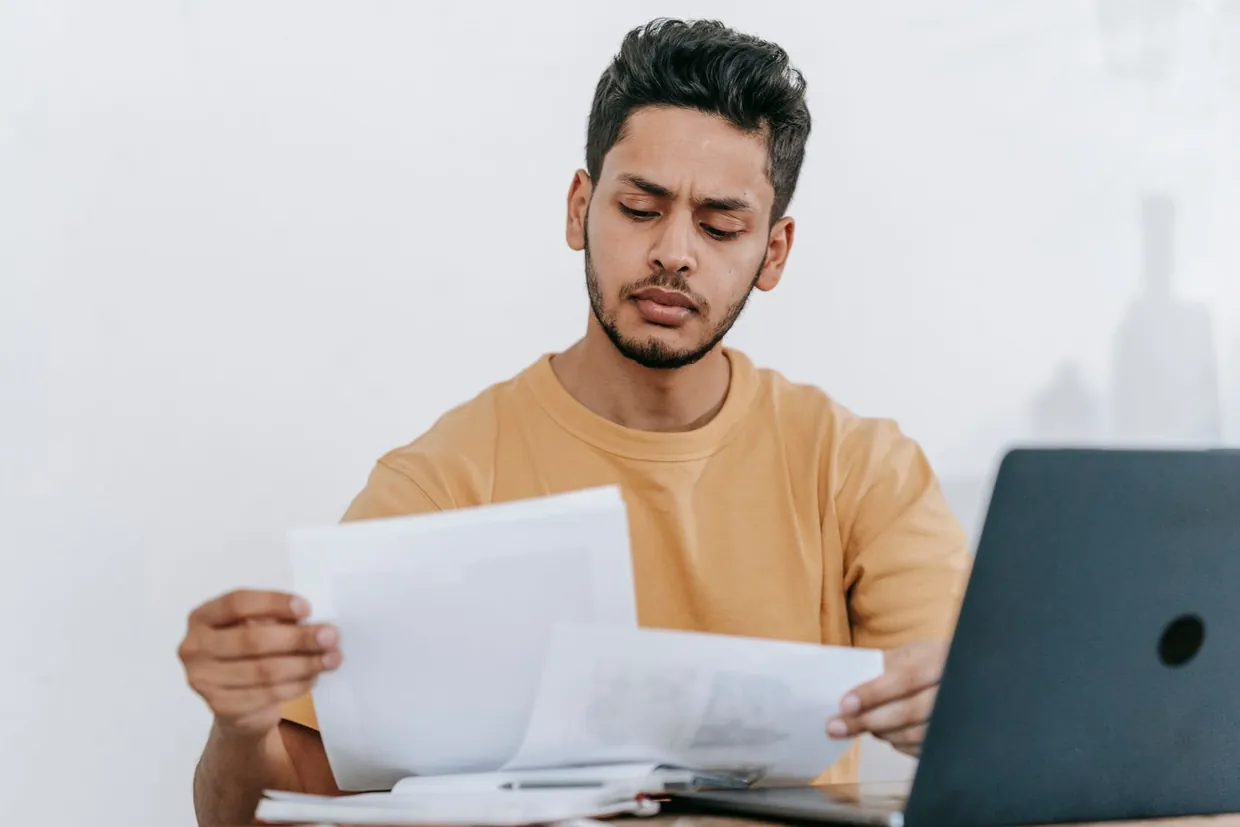 Man comparing two printed proposals at his laptop when costing outsourced customer service